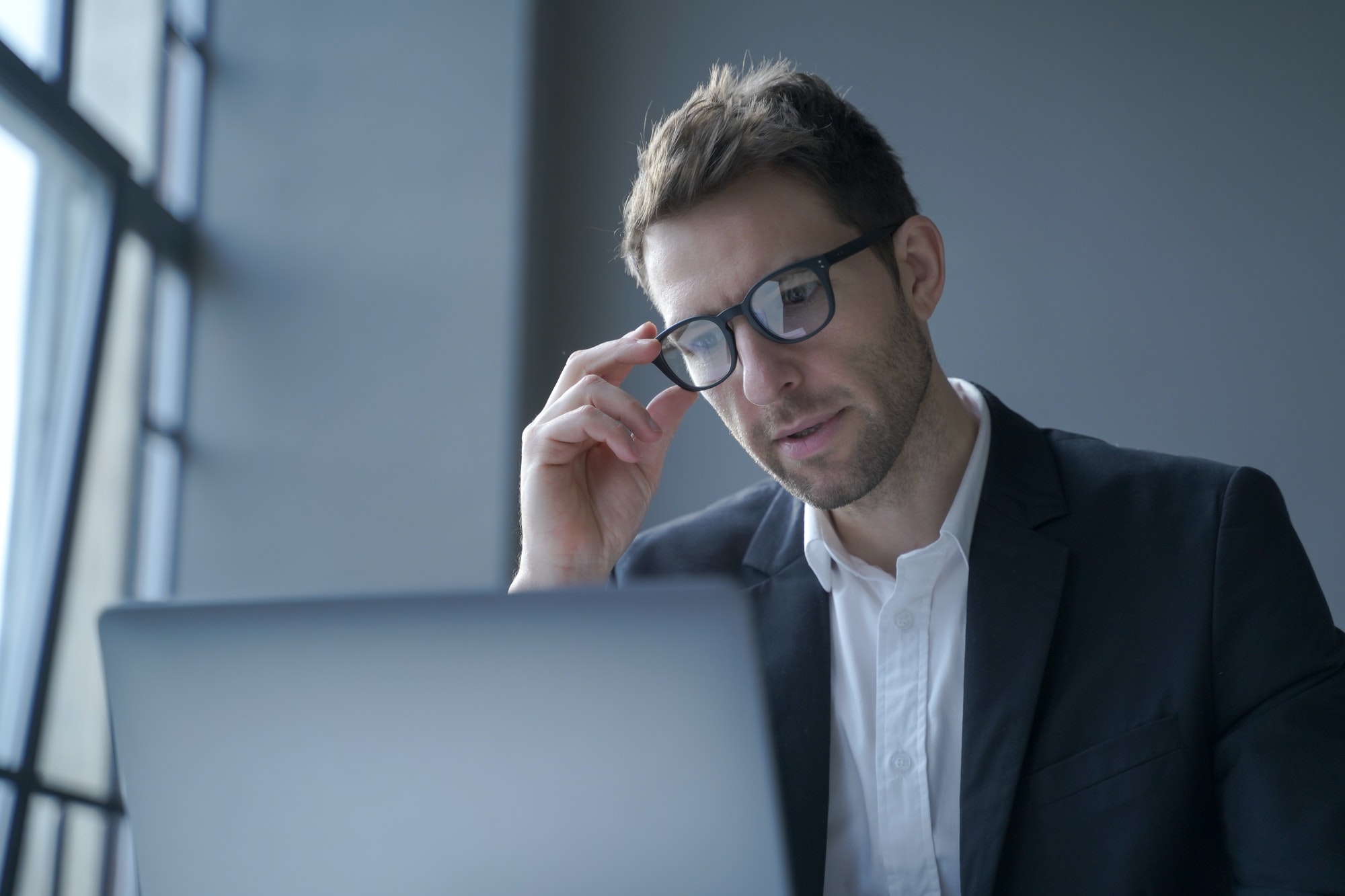 Concentrated young German businessman in formal suit and eyewear looking at laptop screen